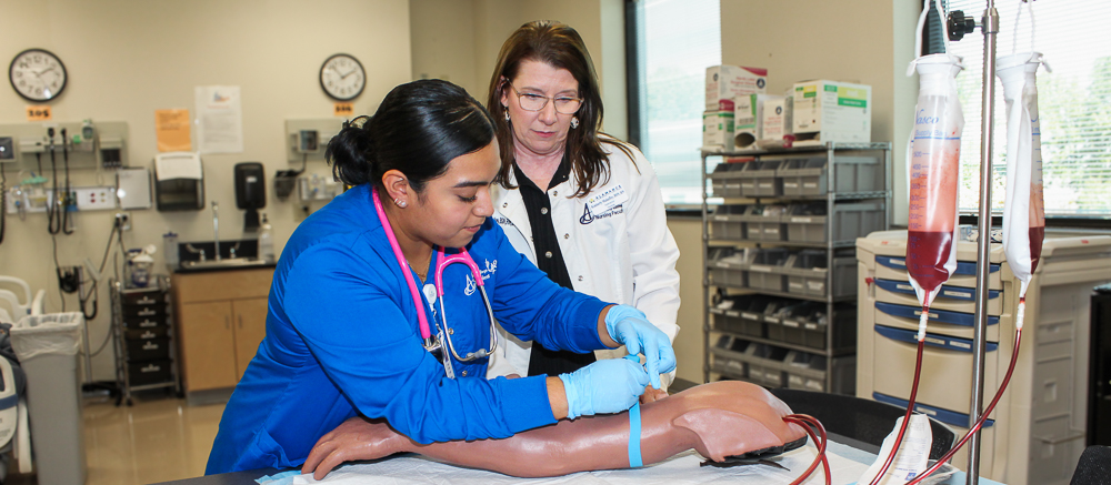 An ACC Phlebotomy student in a blue medical uniform practicing a phlebotomy procedure on a training arm under the supervision of an ACC instructor in a white coat. The setting is a well-equipped medical training lab with IV bags, medical equipment, and shelves in the background, emphasizing a hands-on learning environment