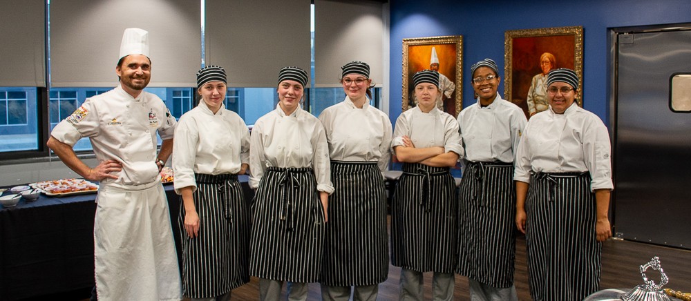 A group of ACC culinary students and their instructor posing together in a professional kitchen or dining space. The students wear white chef coats, striped aprons, and matching hats, while the instructor, in a tall chef's hat, stands to the side. The background includes a blue wall with framed artwork and a table set with food, showcasing a polished and professional setting.