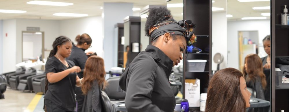 two female cosmetology students working in the acc cosmetology salon. One female in the forground doing hair on a manikin head.