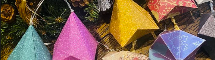 Colorful origami ornaments in various geometric shapes, including pink, blue, yellow, and multicolored designs, displayed on a wooden surface. The background includes green pine branches and pinecones, adding a festive touch